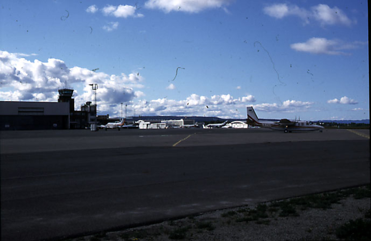 Lufthavn, 1 fly på bakken, GA (Aero) Turbo Comander 690B, LN-FAH fra Fjellanger-Widerøe. Flere fly, kontrolltårnet, hangarbygninger og flyplassbygninger i bakgrunnen.