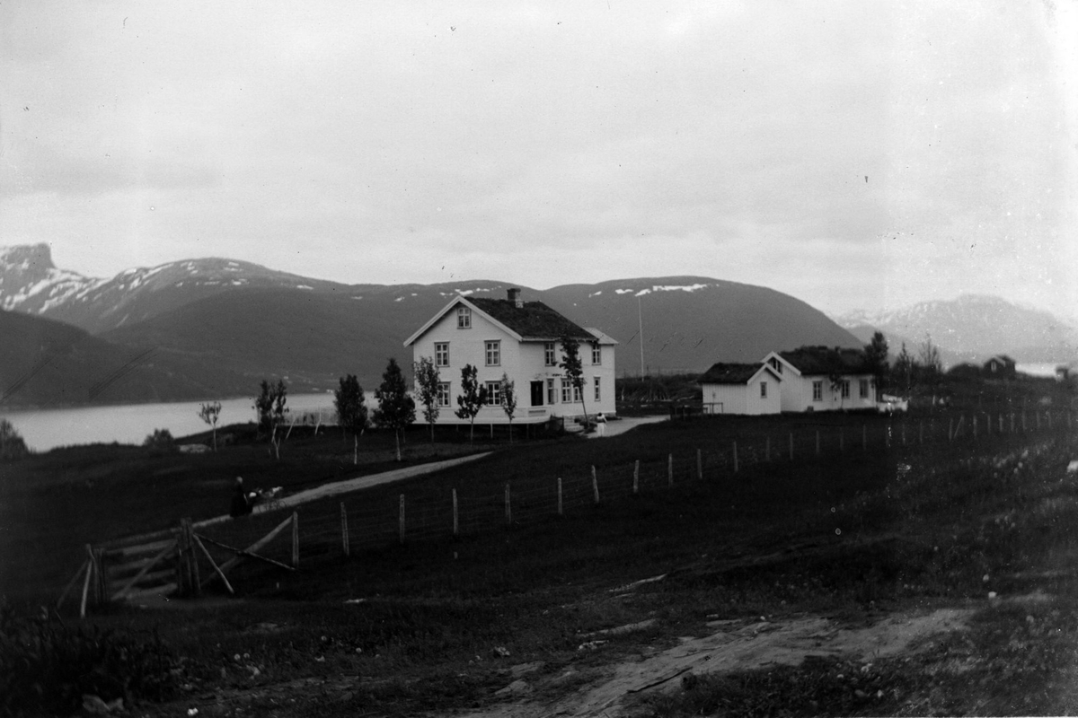 Lensmannsgården i Tysfjord. I forgrunnen en kvinne med barnevogn på veien, fjord og fjell i bakgrunnen.