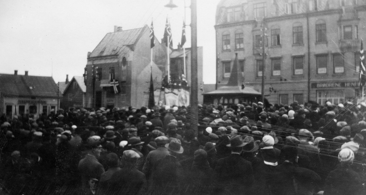 Folkemengde og flagg på Torvet, antagelig på 17. mai. Til høyre i bakgrunnen butikken til brødrene Henriksen.