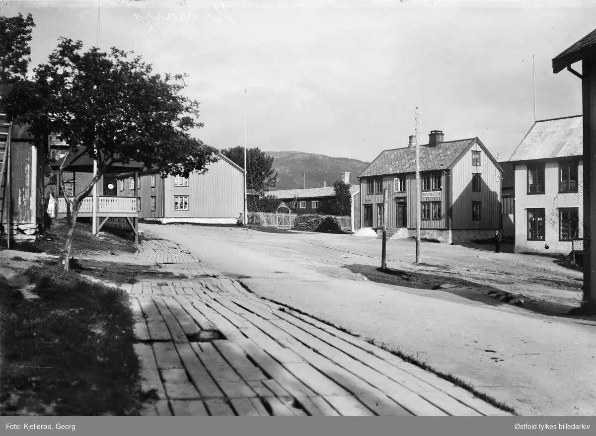 Gateparti fra Hemnesberget i Nordland.  Bygning nr 2 fra høyre har skilt: Rigs Telegraf og Telefon, til venstre lite skilt: Karen(?) Larsen.
Plankebelagt fortau til venstre.