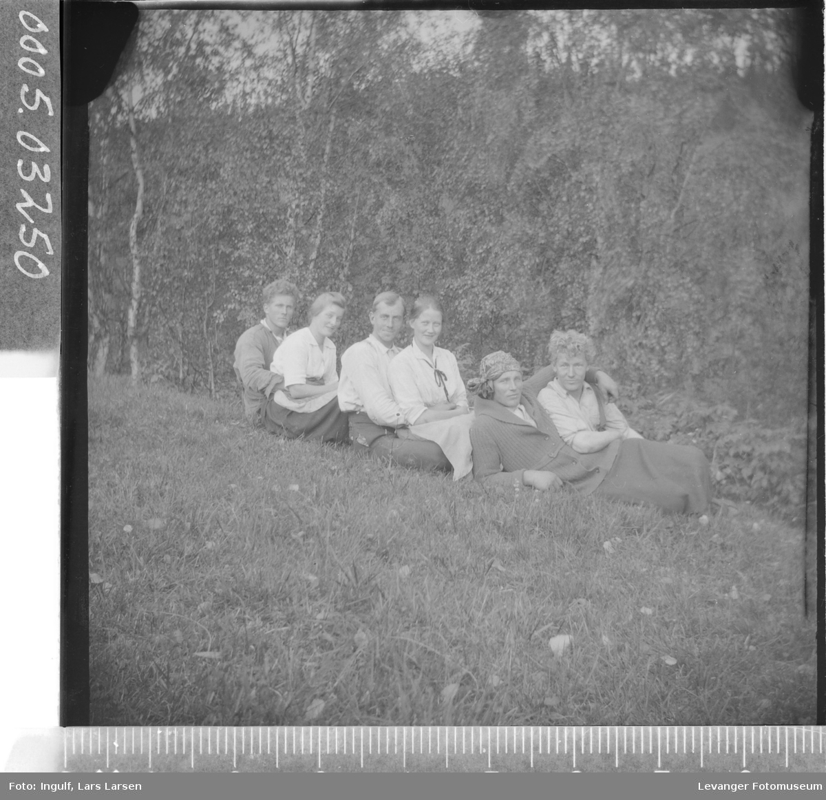 Gruppebilde av fire menn og to kvinner som sitter i gresset, hvorav en mann er utkledd som kvinne.