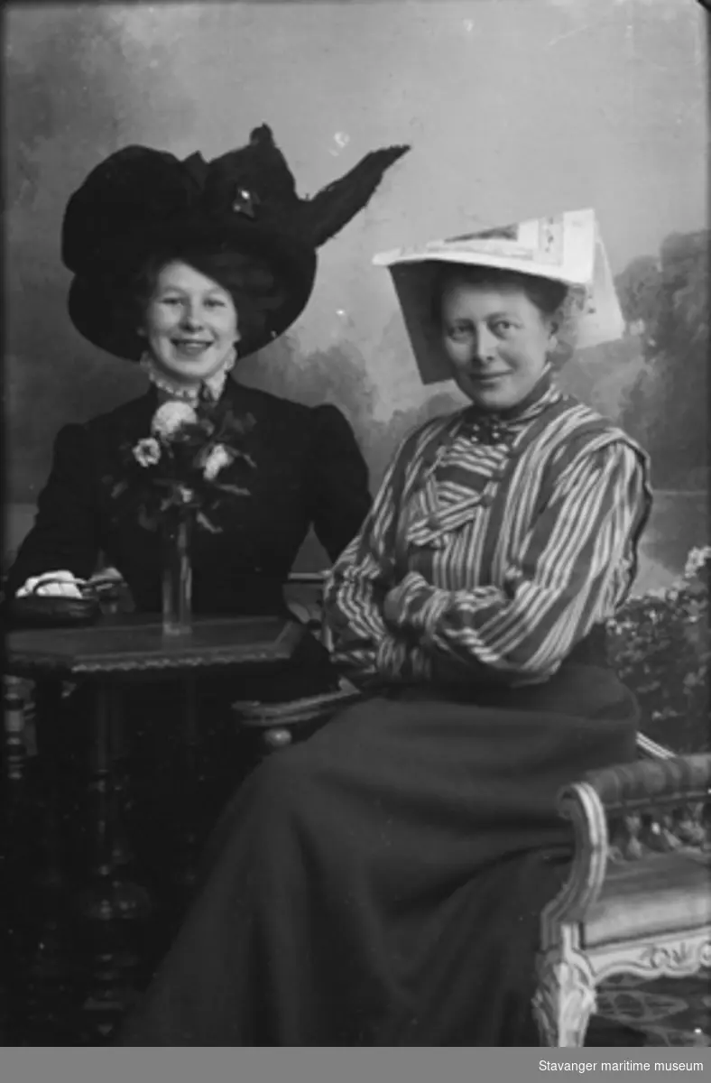 Studioportrett. To kvinner, ukjent ung kvinne til venstre i motivet med stor hatt, blondekrage og pen jakke. Til høyre Julie Lund i stripet bluse og med en avis på hodet. Ca 1905-1910.