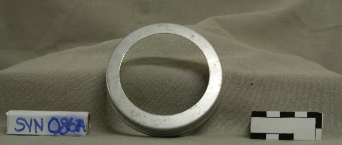 Del 1 er en ring i metall, med riller langs kanten, i ringens høyde. Ringen har skrufeste innvendig. 
Del 2 er en skive i støpt, gjennomsiktig glass, med innskrift på toppen. 