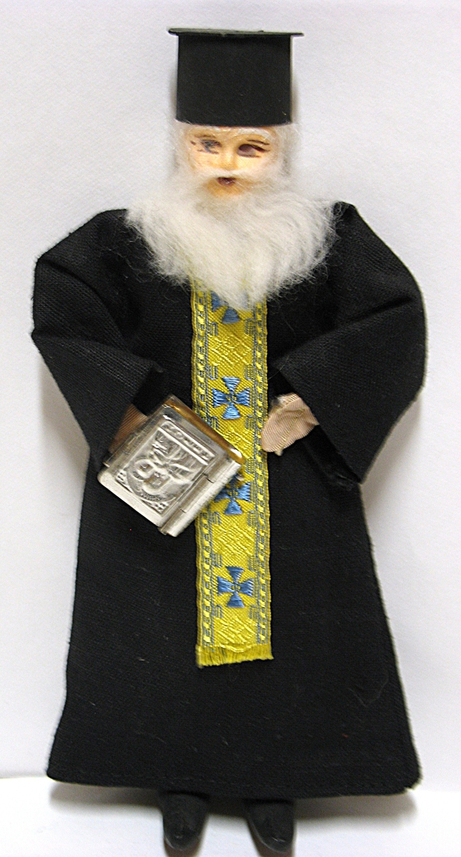 Tillhör Vera Hanssons docksamling.

Prästen Makarios, Grekland. Köpt 1976 på Kreta.
Helt svartklädd. Gult och blått band. Bönebok i handen.