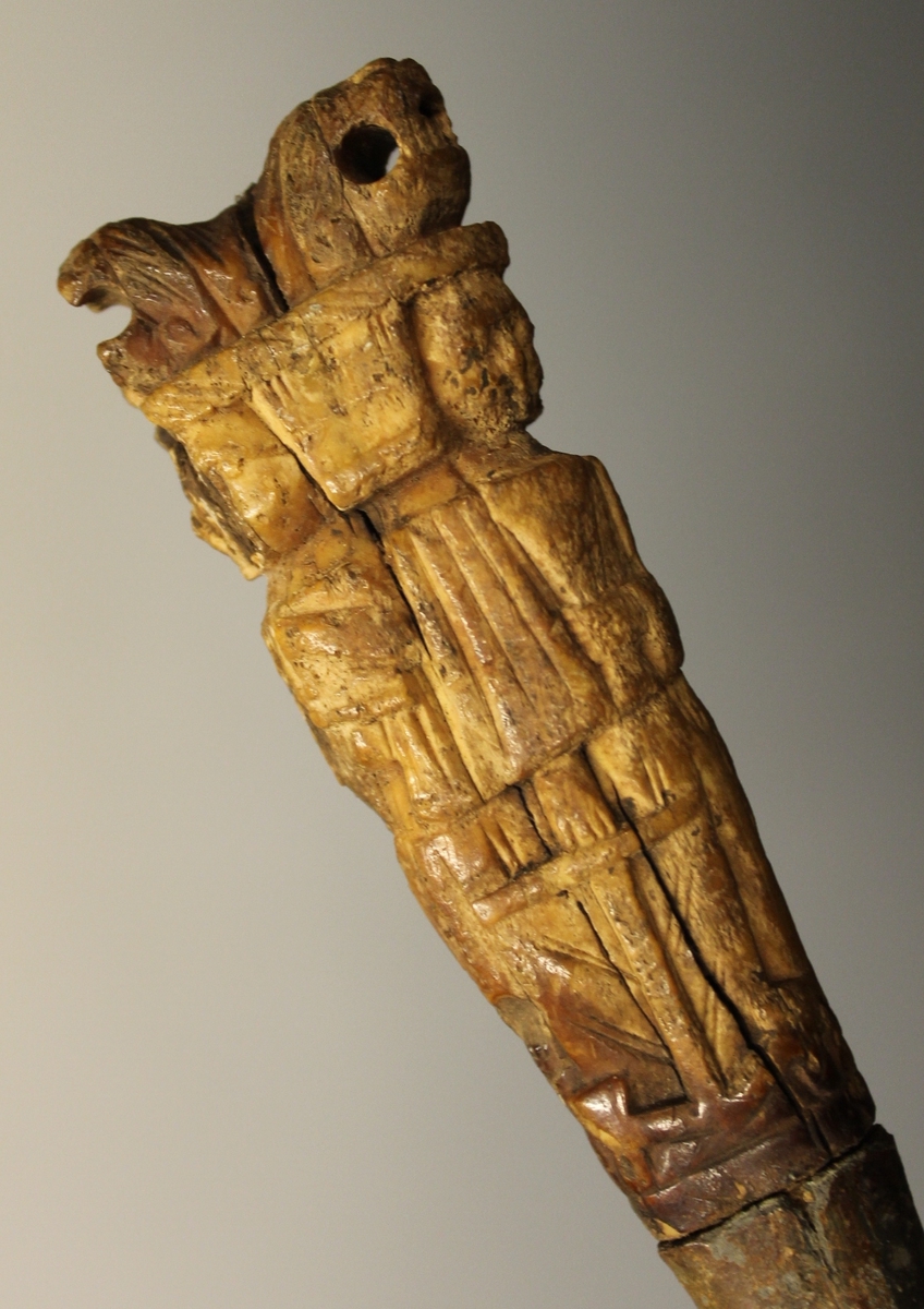 To store figurer hvor en har et  kornaks i hånden, to små figurer - kvinne og mann, anker. Gjennomgående hull i toppen av skaftet. Vanskelig å tyde siden skaftet er slitt og deler brukket av.