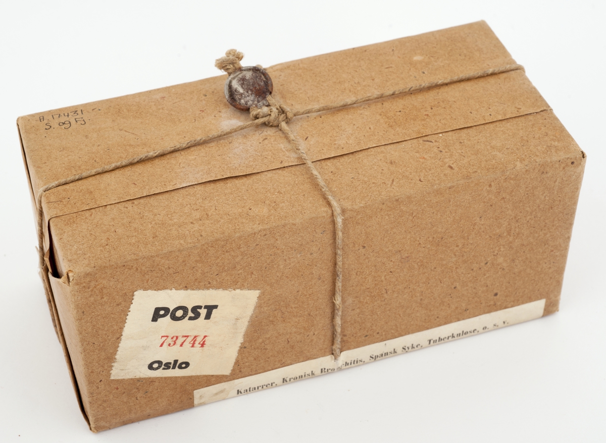 Rektangulær, innpakket postpakke med etikett som både beskriver innhold , produsent og leverandør, og adressant. Uåpnet med hyssing rundt og forsegling. Post-etikett også på pakken.