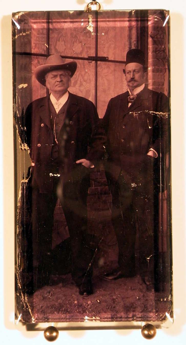 Portrett i helfigur av to middelaldrende herrer med hatt og mørk dress. Fotografiet er tatt utendørs.