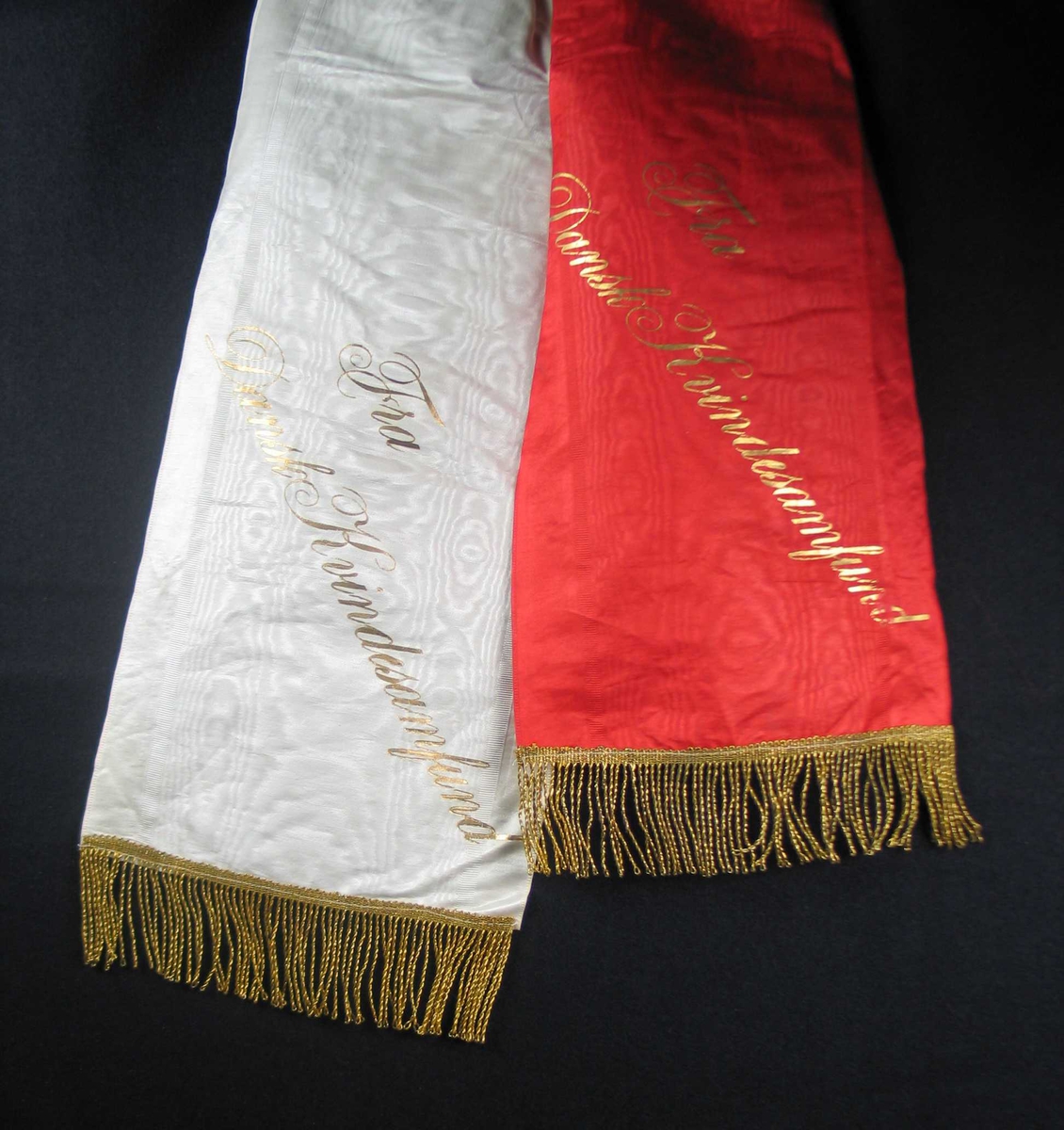 Begravelsessløyfe i rød og hvit silke med frynser og tekst i gull.