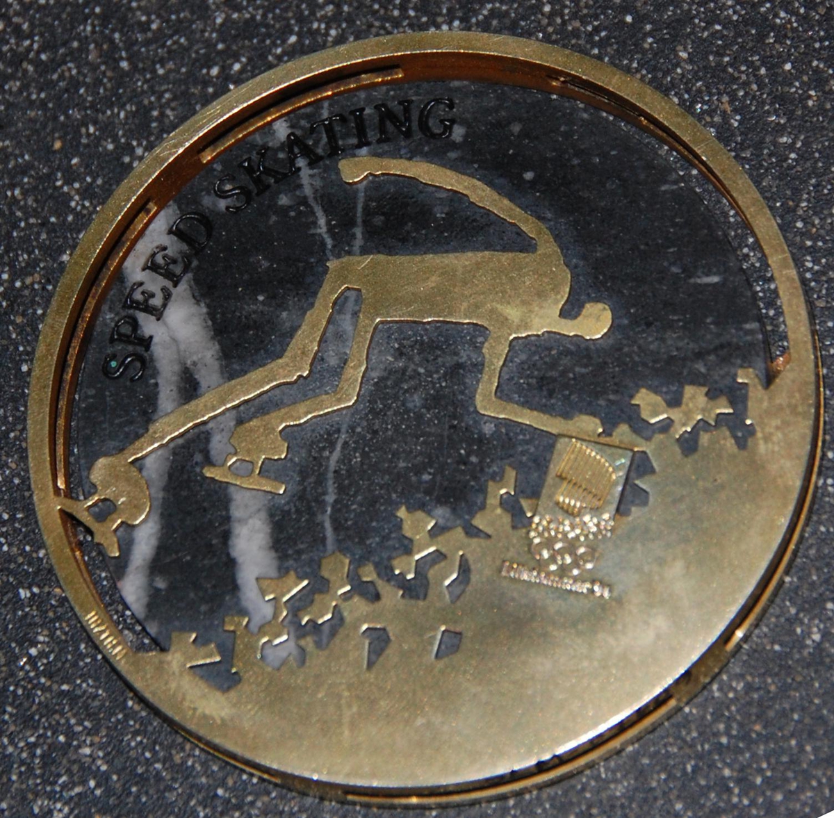 Medalje av metall og stein. På den ene siden er det motiv av de olympiske ringene over krystallmønster. På den andre siden er det piktogram av en skøyteløper og emblemet for de olympiske vinterleker på Lillehammer i 1994. Medaljen er plassert på et stativ av granitt.