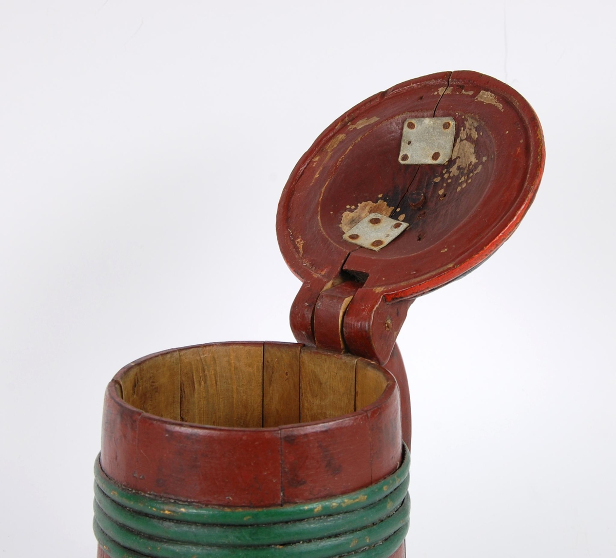 Kanne av furustaver. Lokk med akantusskurd malt i grønne, gule og blåe farger. Kuleformet lokkgrep. Korpus med fire grønnmalte gjorder oppe og en nede (opprinnelig tre). Hanken er utskåret av en av stavene, ender nederst i en plate med bladmotiver.