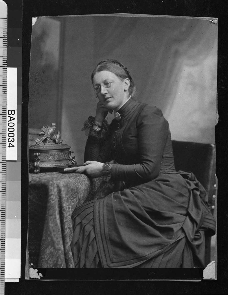 Fotografi av en voksen dame sittende med albuen på et lite bord støttende hode på høyre hånden og med en bok i venstre hånden hvilende på bordet. Hun har briller og oppsatt flette rundt bakhodet. 