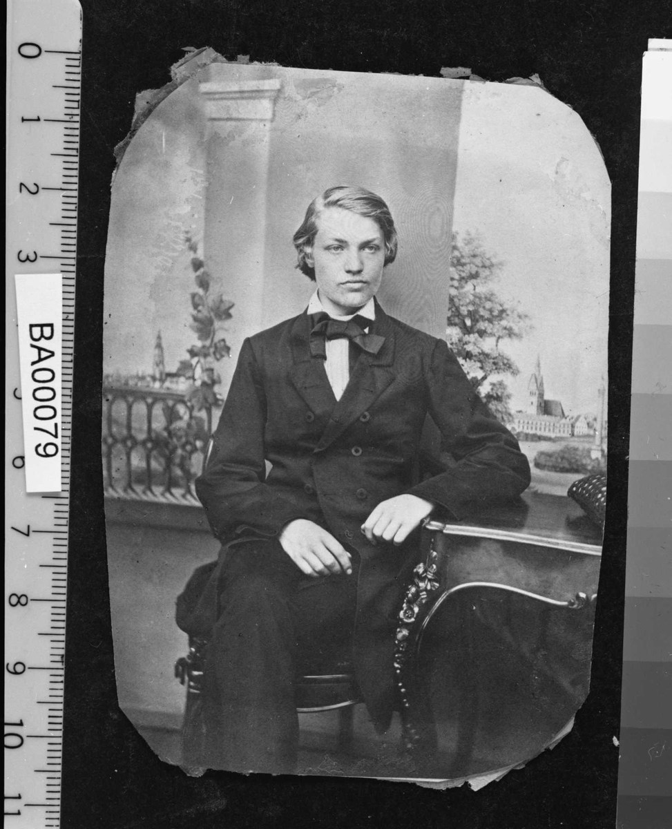 Portrettfotografi av ung mann med halvlangt lyst hår, sittende med benene i kors, kledd i mørk dress med halsbind.