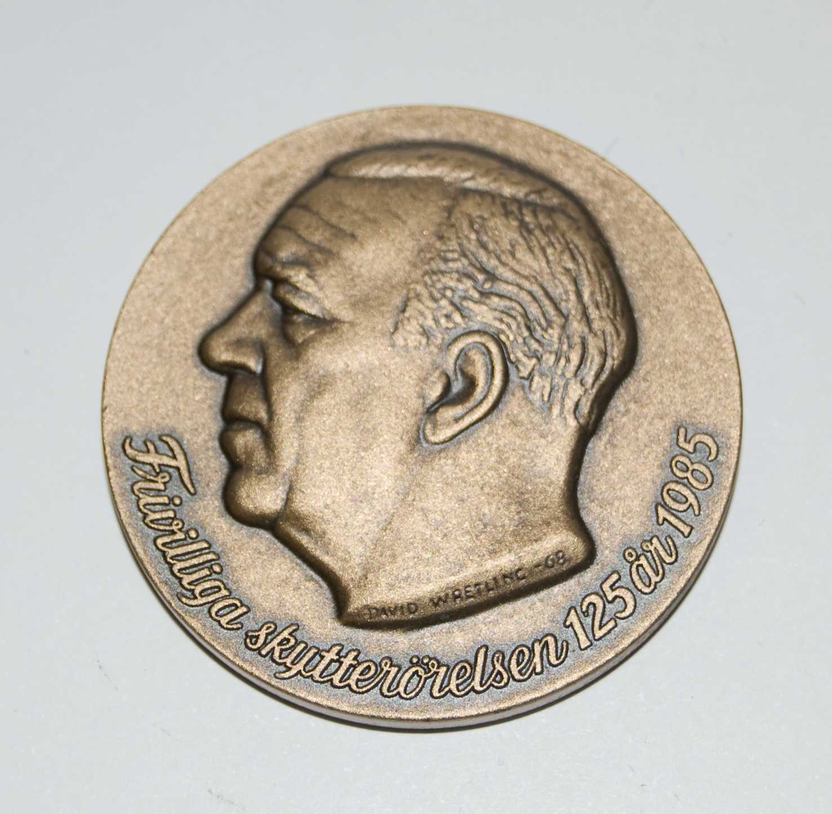 Bronsefarget medalje med motiv av et hode til en ukjent person. Hodet er i profil. Det er også motiv av det som antas å være emblemet til den Frivilliga skytterörelsen.