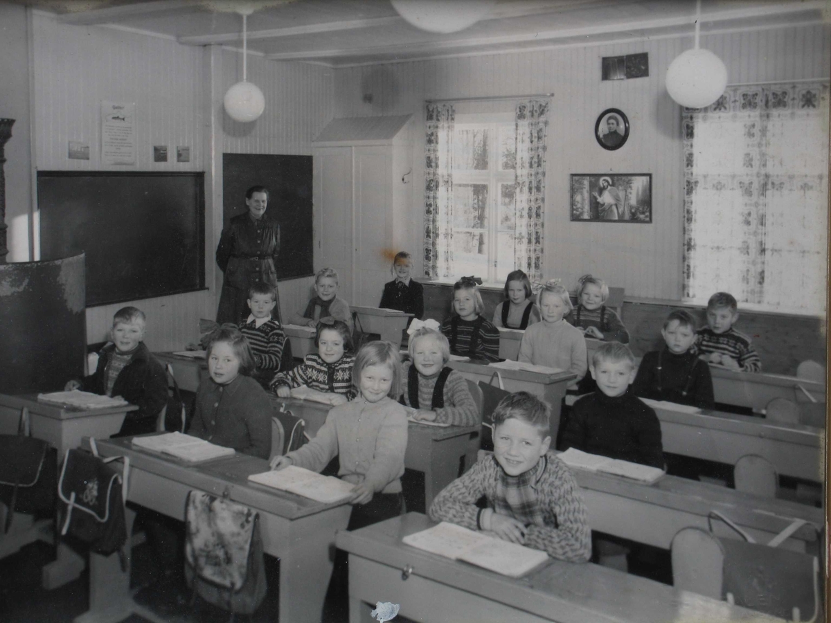 Skoleklasse fotografert i klasserom, elever ved pulter, lærerinne i bakgrunnen. 