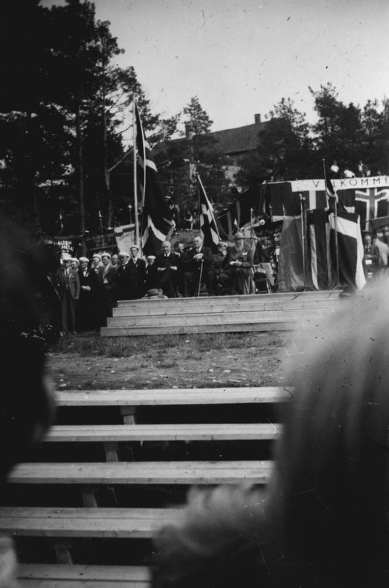 Forsamling ute på en scene med flaggsmykket talerstol. Her sees bl.a. Johan Nygaardsvold.