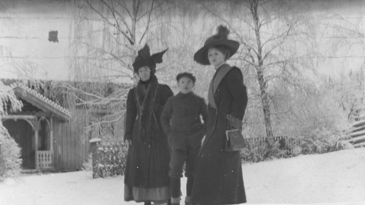 To damer i hatt og kåpe og en gutt i vintersnøen.