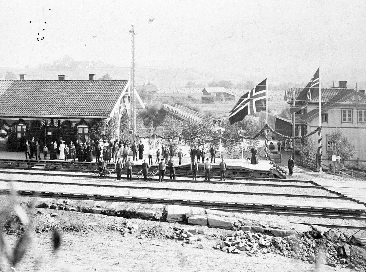 Leirsund Jernbanest. 1. september 1904, ved 50-års jubileet for Hovedbanen. Stasjonen er pyntet med løvranker, og flagg uten unionsmerke. Festkledde mennesker på perrongen. På sporet et arbeidslag og to dressiner.