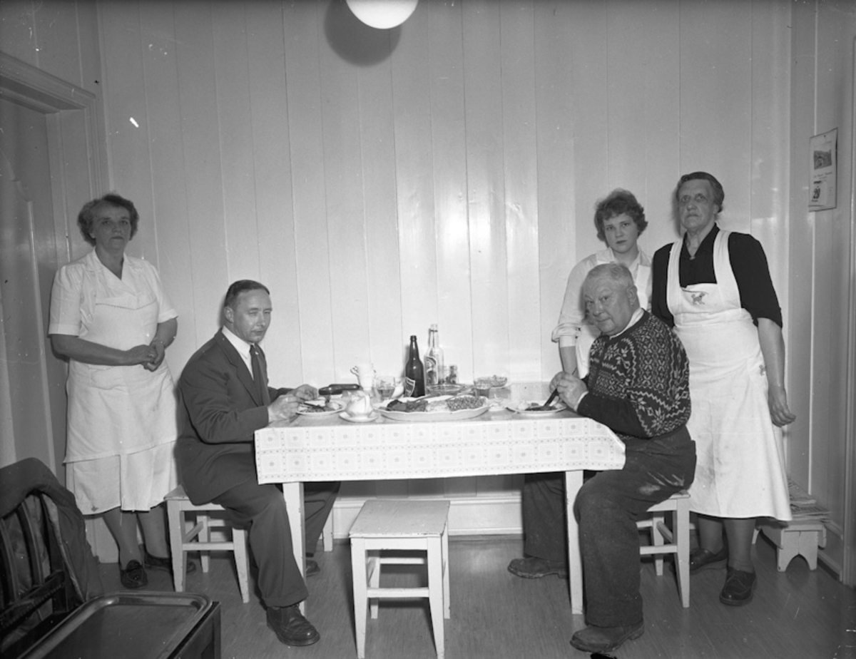 2 menn spiser – 3 kvinner serverer. Øl og akevitt på bordet.