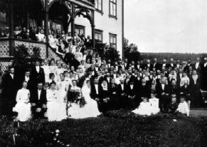 Stor gruppe bryllupsgjester og brudepar på storgård. Nummer tre i første rad fra høyre er Reinert Svendsen.