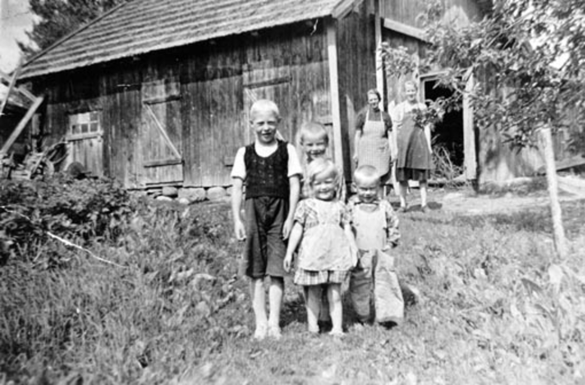4 barn foran uthus. Fra venstre er Magne, Marius, Karen og Hans Myhrhagen. I bakgrunn er Olga Simenstad og Margit Myhrehagen, Nes, Hedmark.