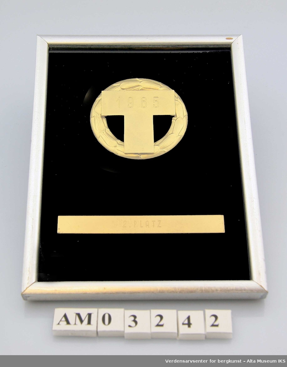 Laurbærkrans m/bokstaven "T" i gullfarget metall på svart bakgrunn. , Rektangulær sølvfarget ramme av tre.