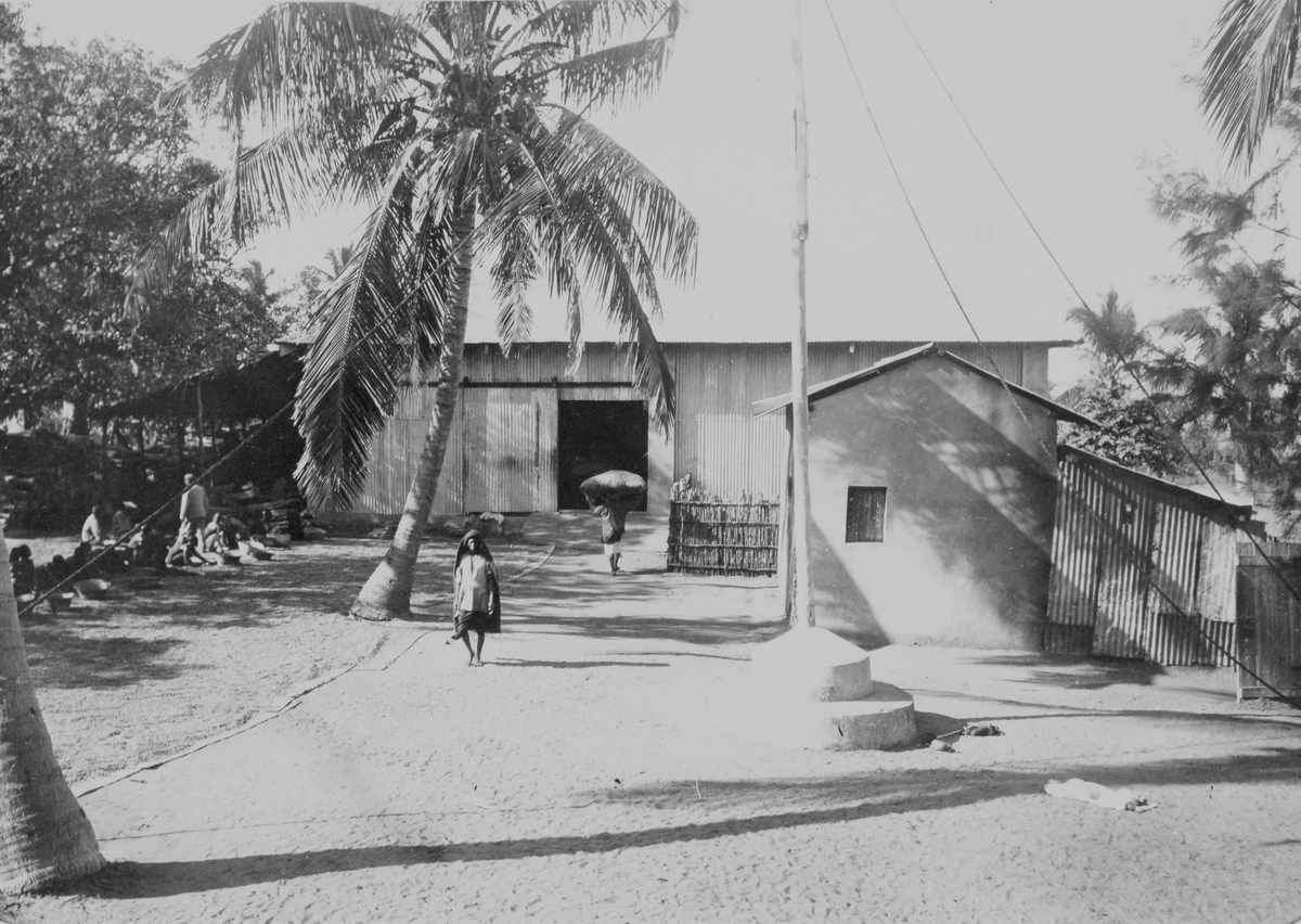 Mosambik. Varehus i bølgeblikk. Arbeidere utenfor. Vakthus, flaggstang og gjerde i bambus. 1914.