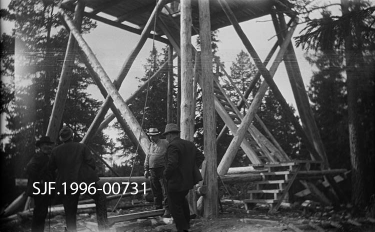 Lundbergseteter skogbrannvaktstasjon fotografert mens tårnet var under bygging 26. februar 1936.  Fotografiet er tatt ved tårnfoten på den åpne, stolpebårne konstruksjonen, der tre dresskledde befaringsdeltakere får en orientering av en busserullkledd tømmermann.  Bildet viser ellers stolpekonstruksjonen med parvise innoverhellende hjørnestolper, krysslagte avstivingsledd og trapp opp til et repos, hvor det antakelig var ny trapp som førte til neste repos, med trapp videre til utkikkshytta på toppen.  I bakgrunnen skimtes glissen gran- og furuskog. 