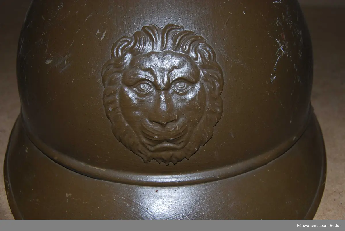 Gulbrun stålhjäm av fransk typ med hakband men utan inredning. Med ett lejonhuvud framtill.
