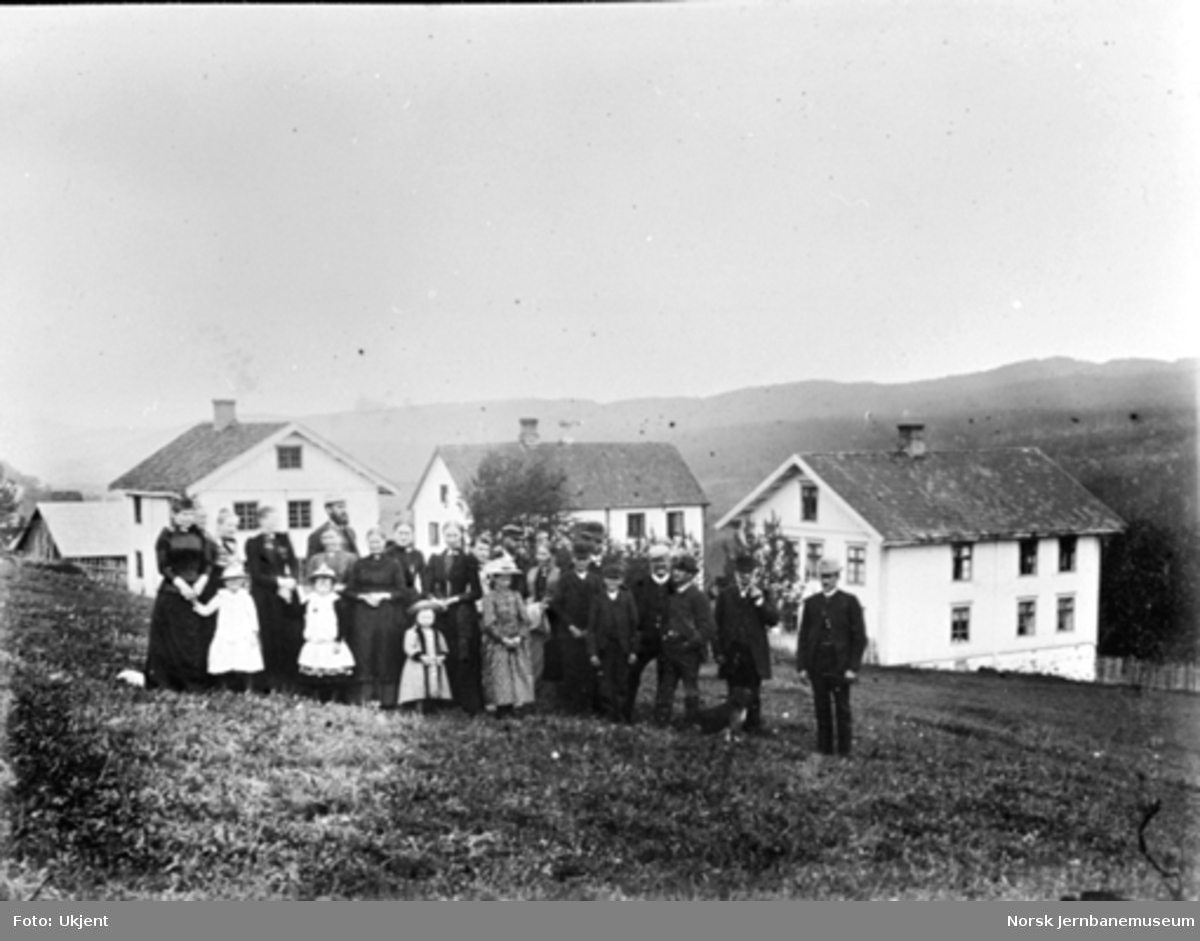 Gruppebilde med 23 personer på gården Stuve i Biri; husene i bakgrunnen