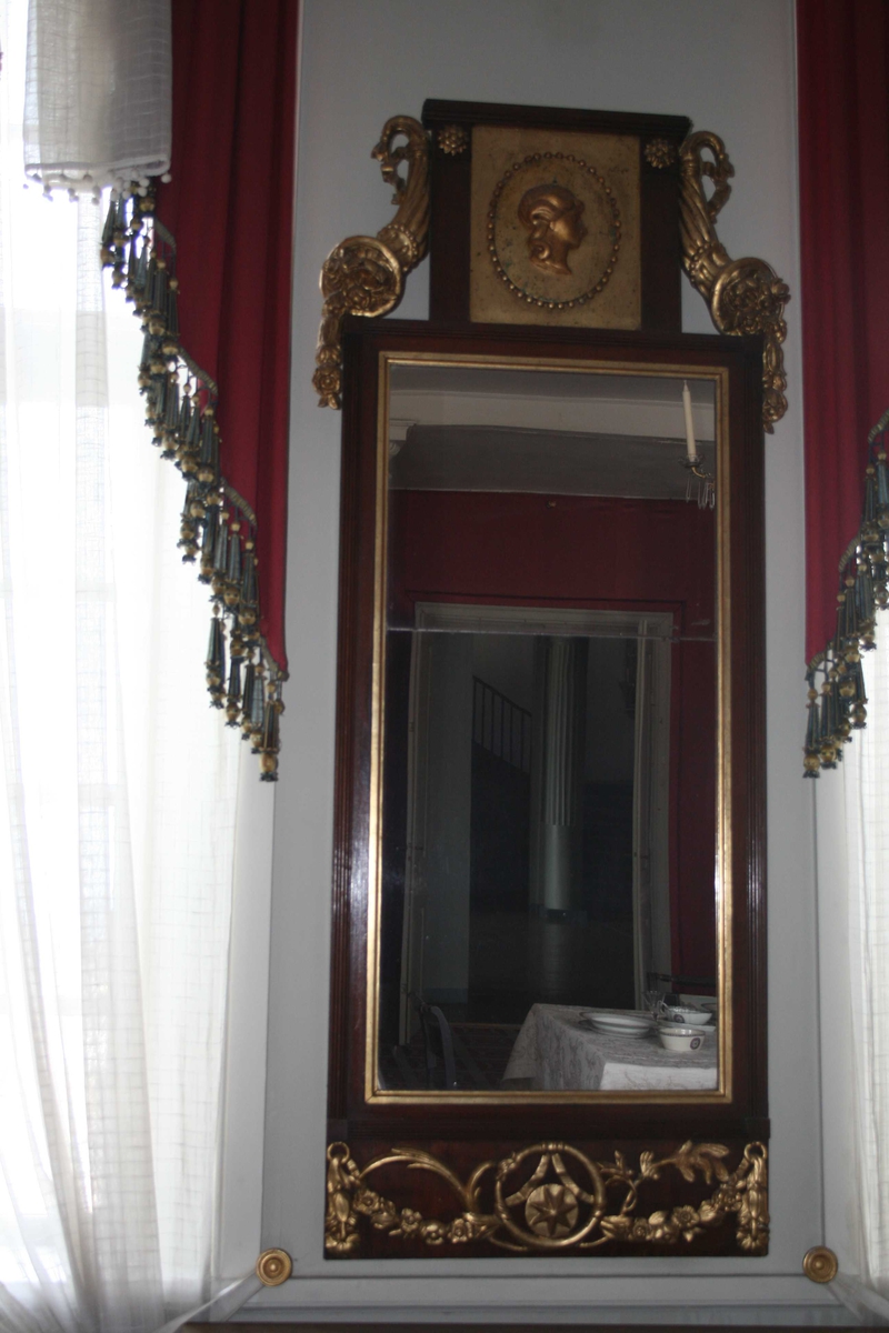 Rektangulært speil med forgylte ornamenter. Toppstykke med medaljong med kvinnehode, overflødighetshorn. Girlandere nederst. Speilglasset er delt i to.