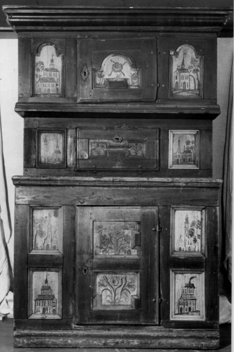 Framskap i en del med en dør nede, en  oppe og en liggende i midten. Dørene har malte blomstermotiver mens de 8 sidefeltene har malte bygninger.