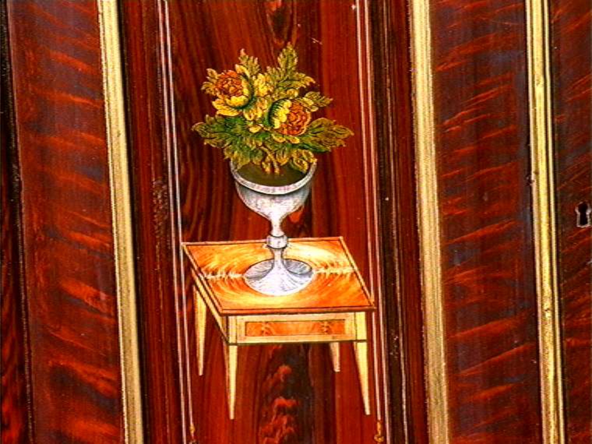 Hjørneskap i to deler. Gullmalte profillister og rosamalt inni. Skapsdørene på overskapet er dekorert med samme motiv på begge dørene, et bord med en vase med blomster.