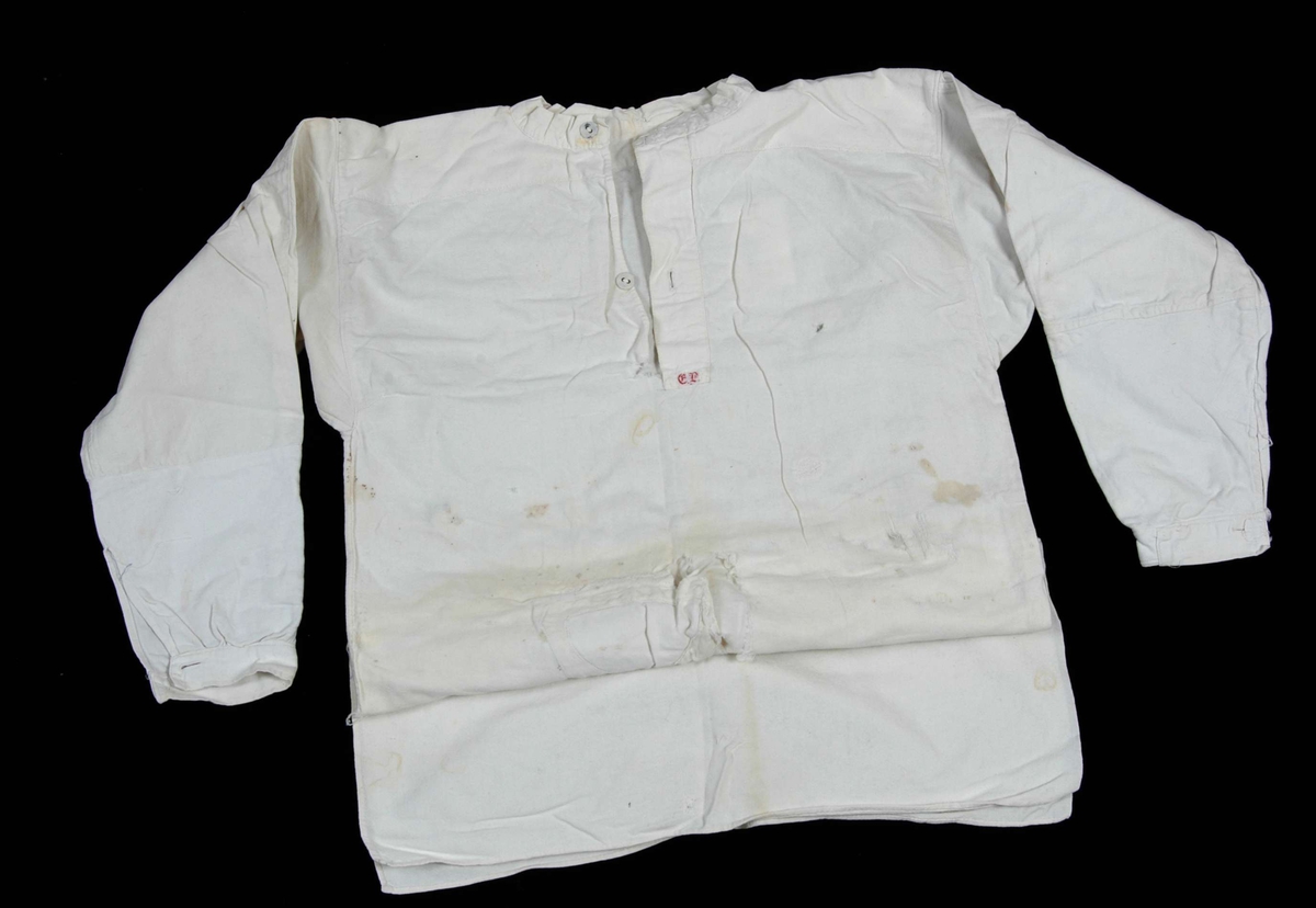 Hvit, lang skjorte med bærestykke, halssplitt med linning og knapp, halslinning med knapp, lange ermer med ermlinning og splitter i sidene. 