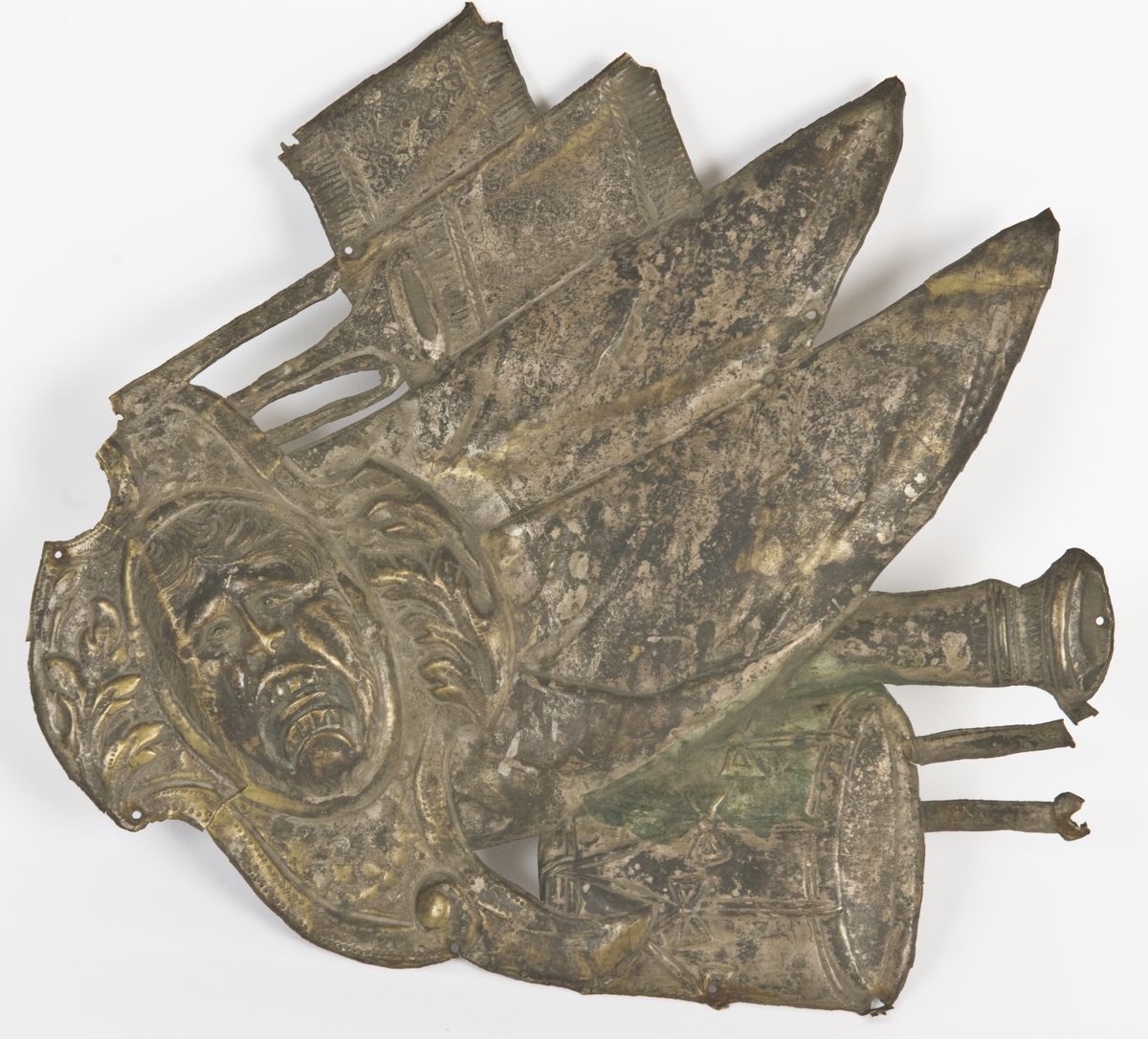 Beslag av hamret messingblikk, iflg. katalogkort fra likkiste. Barokkstil med våpenmotiver (økser, kanoner, trommer, faner etc.) og mannsansikt/maske. Én av tre beslag med samme innføringsnummer fra Nordiska museet (NM.0063777AC).