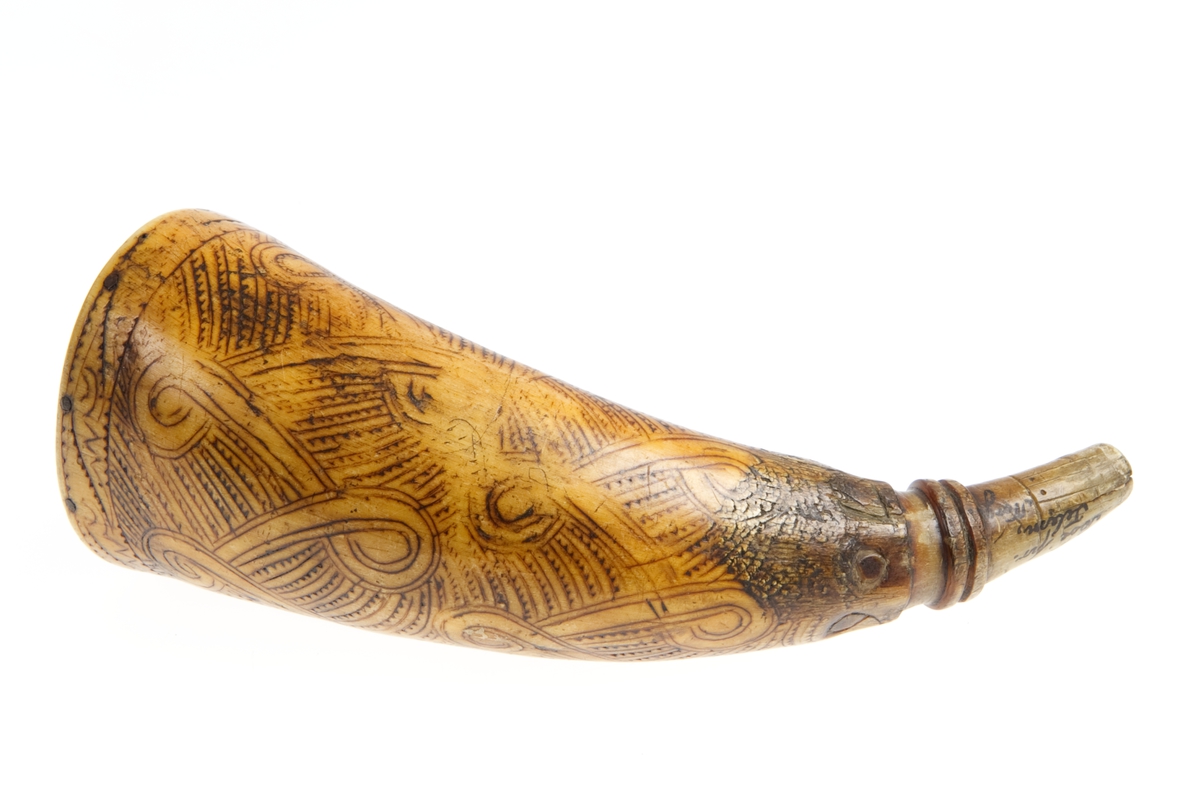 Krutthorn med flat bunnplate i tre med innskrift. Ornamentikk på horn i form av et bånd som går i løkker over hornet mot en bakgrunn av streker med hakk.