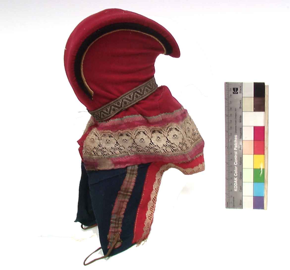 Rød hornlue med med blå og gule detaljer i ull/klede. Dekor av bånd i silke, kniplet hvit bomullsbånd og vevedmetallbånd. Knyttebånd/snor i skinn.
