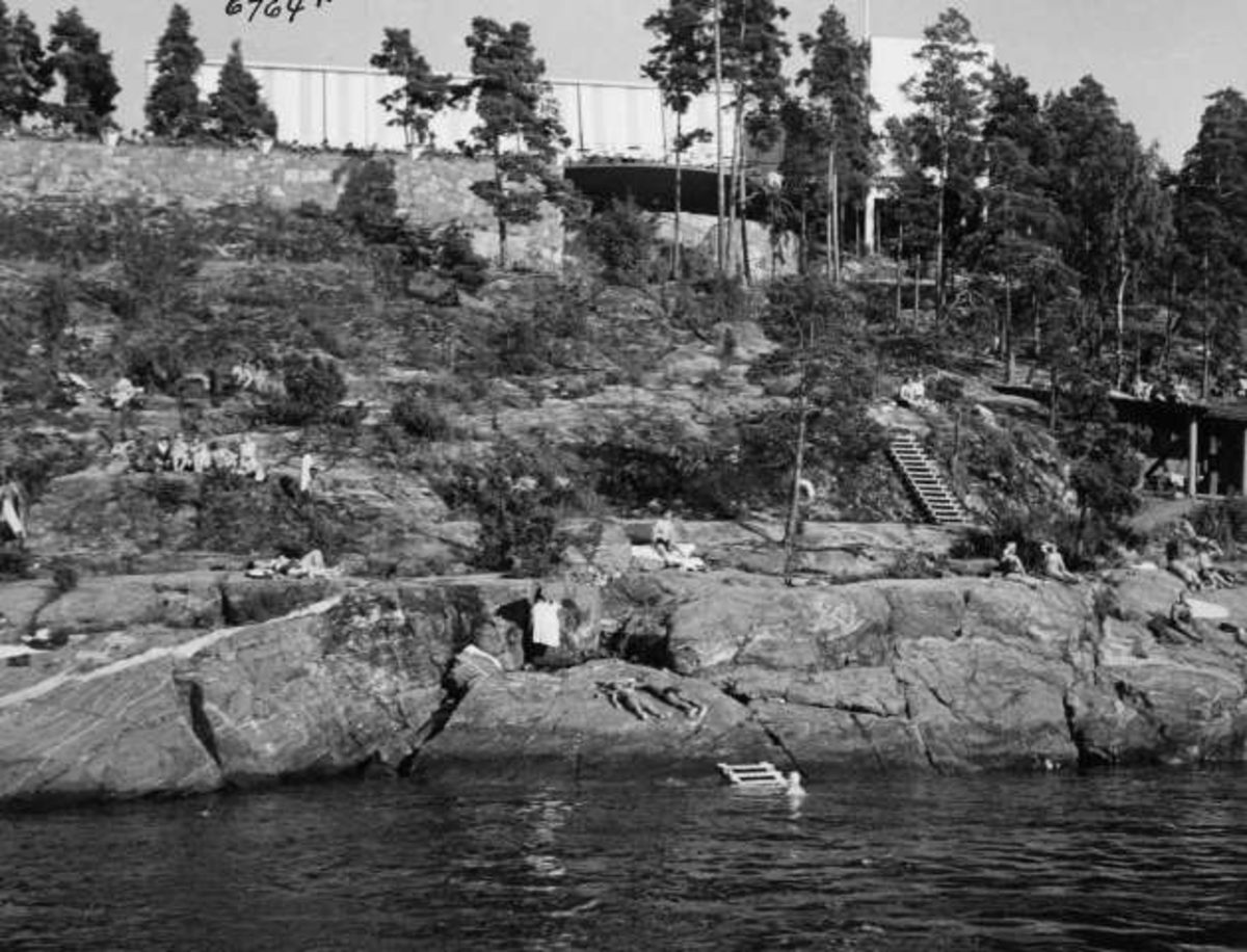 Strandliv med badegjester som soler seg på svaberget ved sjøen på Ingierstrand bad utenfor Oslo. Restauranten i bakgrunnen.