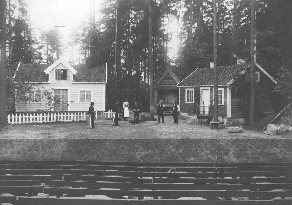 St. Hans feiring på Norsk Folkemuseum med oppsetningen "Friarar" på friluftsteatret. Scene med hus og benkeradene i forgrunnen.