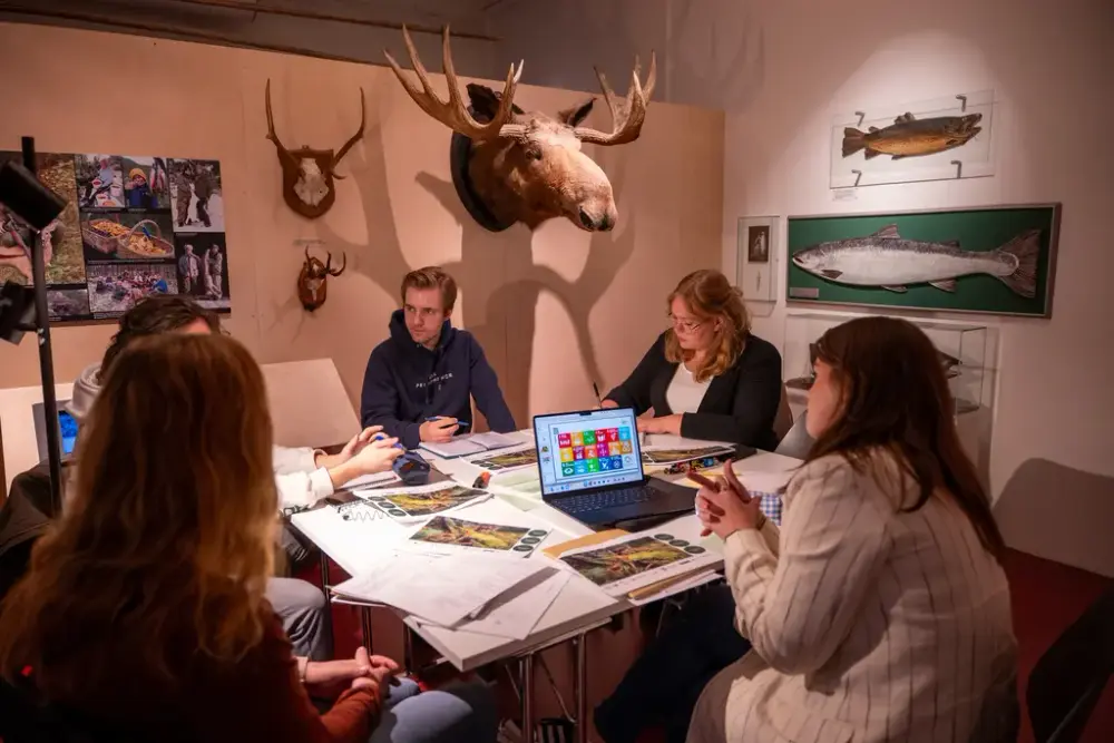 En gruppe ungdommer sitter rundt et bord i utstillingen på Norsk skogmuseum. På bordet ligger det papirer og en pc med FNs mål oppe. I bakgrunnen ser man et elghode og plansjer av fisk.