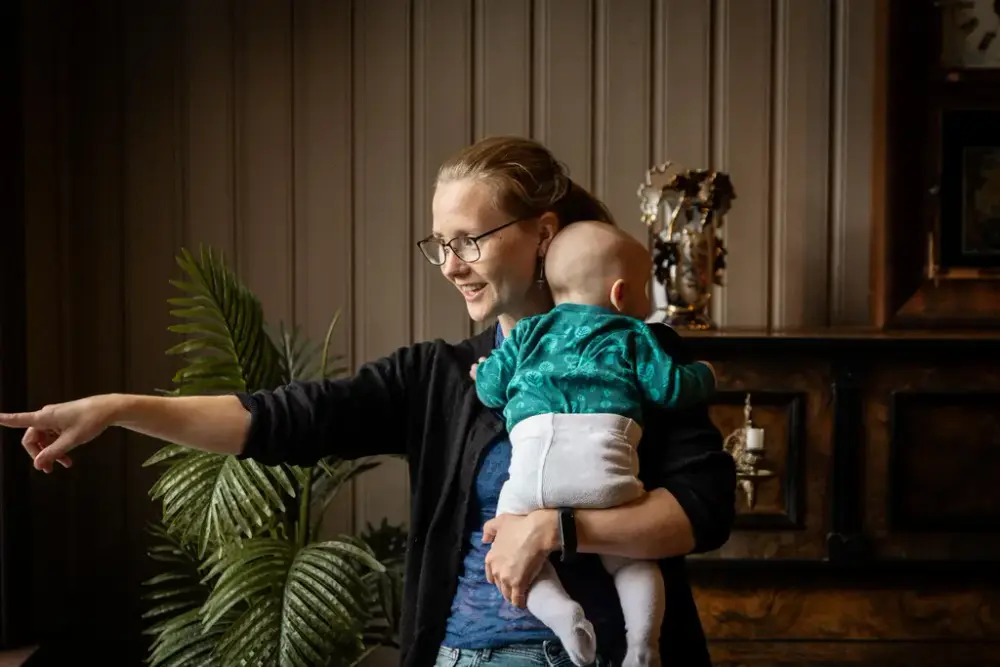 En ung kvinne med en baby på armen peker entusiastisk mot noe utenfor rammen. Bak henne ser vi et gammelt piano i et historisk miljø. 