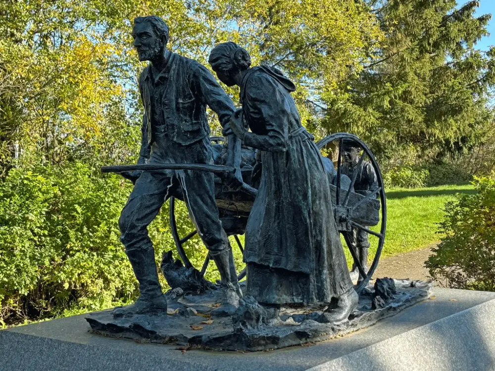 Skulptur av en mann som drar en håndkjerre. En kvinne med lang kjole går ved siden av og et barn går bak kjerra. Skulpturen står på grå sokkel. Gress, busker og trær i bakgrunnen. 