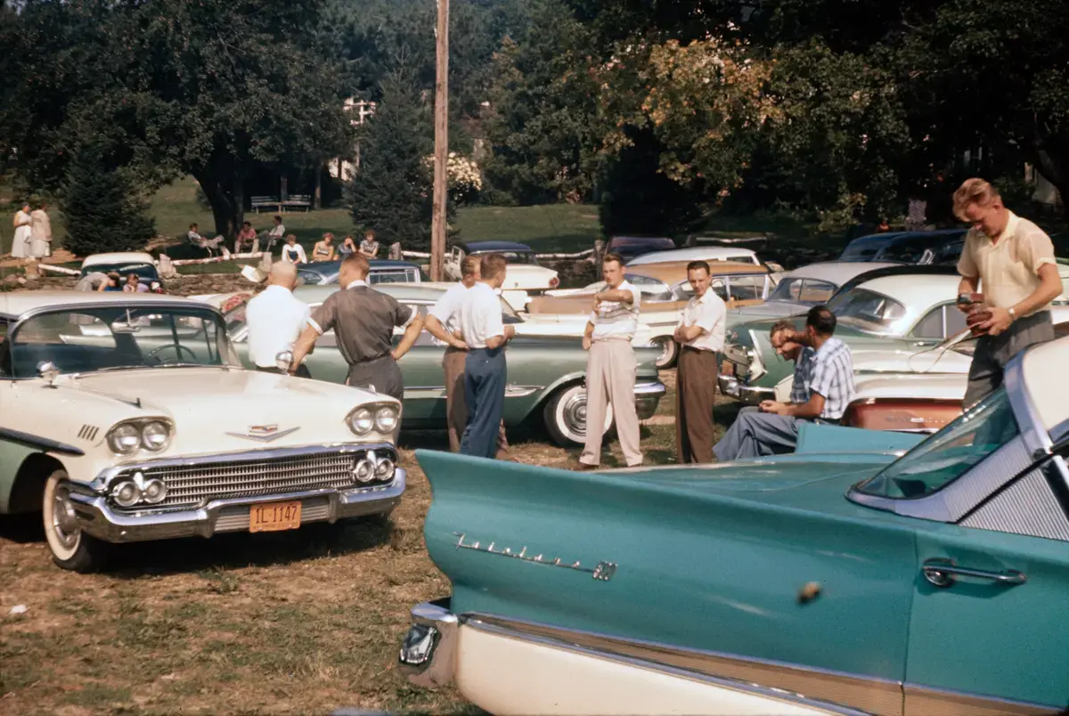 Samling av mennesker rundt amerikanske veteranbiler parkert på gress.